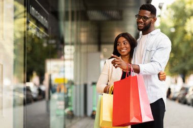 Buraya bak. Gülümseyen Afro-Amerikalı güzel bir kadın moda kıyafetlerini gösteren, mağazanın camını gösteren alışveriş torbalarıyla merkezden geçen, serbest alan olan adama sarılıyor. Aile Alışverişi