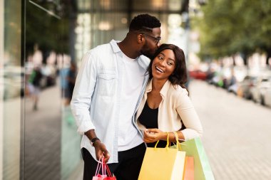 Aşk ve İlişki Konsepti. Romantik siyah çift alışveriş merkezinin penceresinin yanında yürüyor, yakışıklı adam mutlu karısını öpüyor, sevgililer alışveriş torbası tutuyor, geçmişi bulanık.