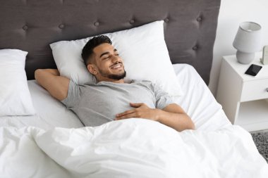 Gülümseyen genç bir Arap, uyandıktan sonra rahat bir şekilde yatağında dinleniyor ve rahat yatak odasının keyfini çıkarıyor. Sabah ışığı etrafındaki neşeli atmosferi güçlendirir..