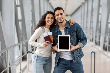 Reklam için Model. Havalimanında poz verirken boş dijital tablet ve pasaport taşıyan mutlu Arap eşleri tasarımınız veya reklamınız için fotokopi alanı gösteren neşeli orta doğu çifti