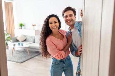 Mutlu genç bir çift modern dairelerinin kapısında duruyor, konukları evlerine davet ediyorlar. Gülüşlerini paylaşıyorlar ve sıcaklıklarını gösteriyorlar. Ziyaretçiler için hoş bir atmosfer yaratıyorlar..