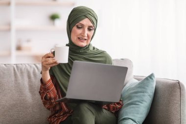 Modern Müslüman kadın dizüstü bilgisayar kullanıyor, sabah kahvesini içiyor, internetten film izliyor ve evdeki koltukta oturmuş haberleri okuyor. Bilgisayar aracılığıyla kıdemli İslami Ev Kadınları Ağı. İnsanlar ve Gadget