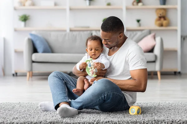 Babalık kavramı. Mutlu siyah adam, sevimli küçük çocuğuyla evde oynuyor, Afrikalı Amerikalı babasıyla ilgileniyor ve küçük bebek kucağında sevimli çocuk oyuncaklarıyla vakit geçiriyor.