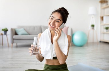 Genç, zinde bir Hintli kadın bir bardak su tutuyor, akıllı telefondan konuşuyor, ev içi eğitimden kaçıyor. Neşeli milenyum doğulu bayan ev çalışmasından sonra telefon konuşmasının keyfini çıkarıyor.