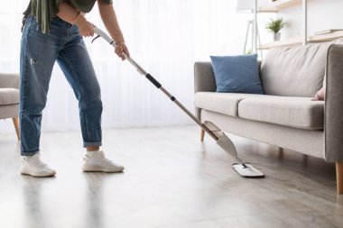 Ev işleri konsepti. Kapalı kesim, başörtüsü takmış, başörtülü, İslami kadının, kanepenin altındaki ahşap döşemeleri, su spreyi ve temizleme maddesiyle doldurulabilir şişeyi kullanarak yıkıyor.