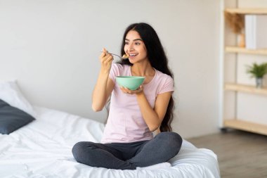 Çekici Hintli kadın evde rahat bir yatakta otururken sütlü mısır gevreği yiyor. Genç Doğulu kadın sağlıklı yemek yiyor, dengeli kilo verme diyeti uyguluyor.