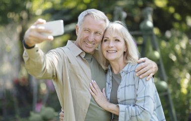 Mutlu yaşlı bir çift bahçede birlikte vakit geçiriyorlar, selfie çekiyorlar ve cep telefonu kameralarına gülümsüyorlar. Mutlu emeklilik ve yaşlı yaşam tarzı konsepti