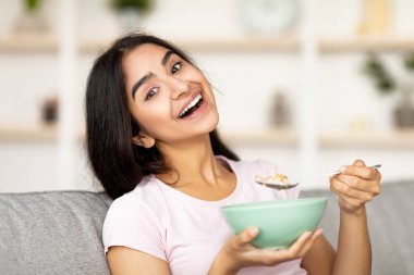 Evdeki rahat koltukta gevrek ve süt yiyen neşeli Hintli kadın kameraya gülümsüyor. Sağlıklı yaşam tarzını sürdüren, sağlıklı yemek yiyen güzel bir doğu kadını. Dengeli beslenme kavramı