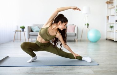 Güzel Hintli bir kadının uzun bir portresi vücudunu esnetiyor yoga minderinin üzerine eğiliyor ve evdeki karantina sırasında aktif yaşam tarzına öncülük ediyor. Güzel Asyalı kadın içeride çalışıyor.