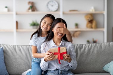 Güzel Asyalı kız, annesi için doğum günü sürprizi hazırlıyor ve oturma odasındaki kanepede otururken annesinin gözlerini kapıyor. Mutlu kadın hediye kutusunu tutuyor.