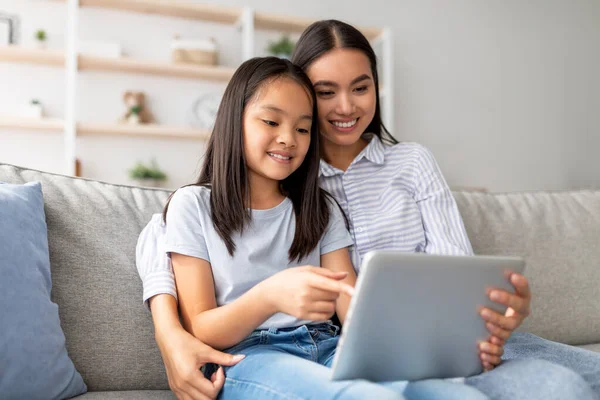 Heyecanlı Asyalı anne ve kızı kanepeye oturup internette sörf yaparken dijital tablet kullanıyorlar. Mutlu anne ve çocuk internette oyun oynuyor ve sarılıyorlar.