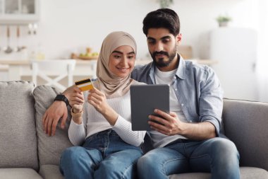 Covid-19 karantina, hızlı alışverişler, evde birlikte alışveriş. Mutlu bin yıllık Arap kadın tesettürlü ve kocası tablete bakar, oturma odasındaki kanepede kredi kartı faturasını öder.
