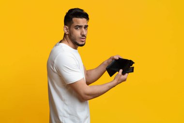 Financial crisis and bankruptcy while COVID-19 pandemic. Unhappy sad middle eastern millennial man showing empty wallet at camera, short of money, yellow studio background, copy space
