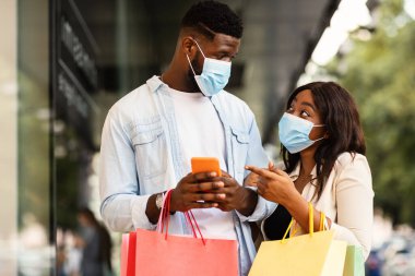 Perakende satış, Teknoloji, Koruma ve Salgın Konsepti. Ameliyat maskesi takan, cep telefonu kullanan, ekranı gösteren, alışveriş torbası tutan, mağazanın yanında yürüyen heyecanlı siyahi çiftin portresi.