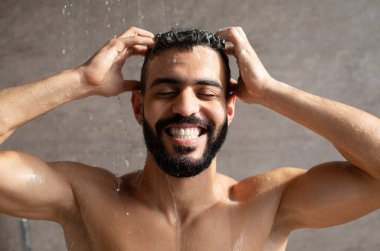 Yakışıklı genç Arap adam duşta duruyor, saçına şampuan sürüyor. Kafasına masaj yaparken gözleri kapalı gülümsüyor..