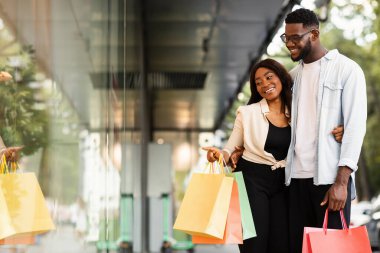 Hadi bunu alalım. Gülümseyen Afro-Amerikan bir çiftin portresi alışveriş merkezinin yakınında geziniyor, vitrinlere bakıyor, kadın alışveriş merkezinin penceresini işaret ediyor. Mevsimlik Satış ve İndirim, Özel Teklif