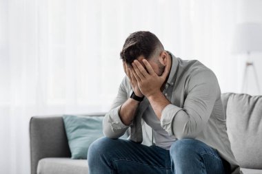 Utanç verici umutsuzluk, üzgün adamın portresi baş ağrısı, stres ve depresyondan muzdarip. Bin yıllık mutsuz Avrupalı yakışıklı adam ağlıyor, yüzünü ellerle kapatıyor, oturma odasındaki kanepede oturuyor.