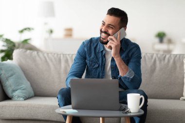 Evden çalışıyorum. Kapalı mekanda dizüstü bilgisayarla çalışırken cep telefonuyla konuşan yakışıklı genç bir Arap. Milenyum Doğulu adam uzak iş ve mobil iletişimin keyfini çıkarıyor