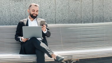 Takım elbiseli, dizüstü bilgisayarlı, ofisin yanındaki bankta oturan, akıllı telefon kullanan, müşterilerle sohbet eden, boş alan olan bir panorama. Modern laptop ve cep telefonu kullanan kendine güvenen bir adam.