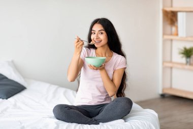 Evde rahat bir yatakta otururken mısır gevreğiyle süt ya da mısır gevreği yiyen çekici Hintli kadın. Güzel Asyalı kadın sağlıklı yemek yiyor, dengeli besleniyor.