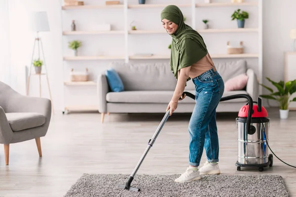 Oda servisi konsepti. Süpürge kullanarak başörtüsü takan genç müslüman kadın oturma odasında ya da yatak odasında gri halı yıkıyor. Bulanık Arkaplan, Seçici Odaklanma, Boş Kopyalama Boşluğu