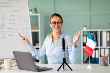 Yabancı bir dil öğretmeni akıllı telefon ve dizüstü bilgisayarla çevrimiçi olarak Fransızca öğretiyor. O, parlak, düzenli bir çalışma alanında öğrencilere dil bilgisi kurallarını özgüvenle açıklıyor..