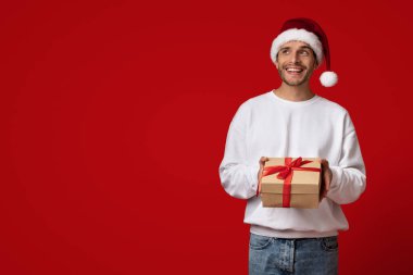 Noel Baba şapkalı neşeli bir adam parlak kırmızı bir arka fonda duruyor, elinde güzel paketlenmiş kırmızı kurdeleli bir hediye kutusu, bayram sezonunu kutluyor..