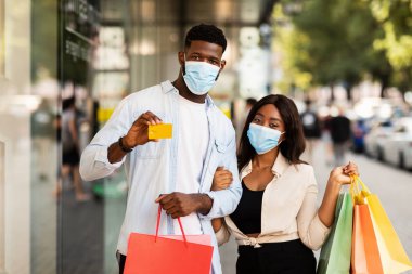 Kolay ödeme. Cerrahi yüz maskesi takmış siyah bir çiftin portresi ellerinde alışveriş torbaları olan altın bankamatik kartlarını gösteriyor, alışveriş merkezinin yakınından geçiyorlar, seçici odaklanma, bulanık bir arka plan.