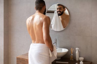 Genç bir Arap adam şık bir banyoda üstsüz duruyor. Yuvarlak aynaya bakarken yüzünü havluyla siliyor. Cilt bakımında mutlu ve kendinden emin görünüyor..