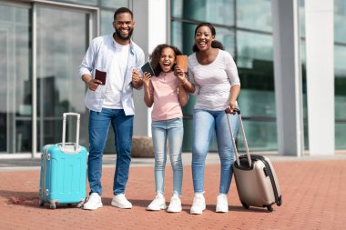 Sonunda tatil zamanı. Biletleri olan ve yumruklarını sıkmış üç kişilik neşeli, pozitif siyah aile, kazanan jest yapıyor, havaalanı terminalinin yanında birlikte duruyorlar.