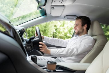 Rahat bir kişisel ulaşım, yeni bir araba, iş adamı ofise gider, yüz ifadesi pozitiftir. Beyaz gömlekli neşeli beyaz adam direksiyonda oturur ve şehirde kahve ile gezer.