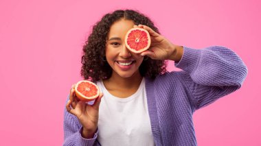 Bir kadın her iki elinde bir greyfurtun iki yarısını tutarken gülümser. Beyaz bir gömlek ve mor bir kazak giyiyor. Arka plan parlak pembe, neşeli bir ortam yaratıyor..