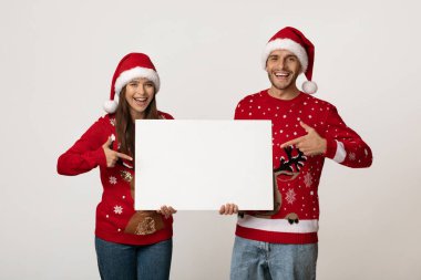 Neşeli bir çift Noel sezonunu Noel süveterleri ve Noel Baba şapkalarıyla kutluyor. Gülümsüyor ve boş bir tabelayı işaret ediyorlar. Mesajlara ya da selamlara hazırlar..
