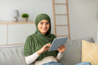 Yeşil tesettürlü neşeli genç bir Arap kadın rahat bir koltukta oturup dijital tablet kullanıyor ya da çalışıyor. İnternete girip evde dinlenirken internete bağlanıyor..