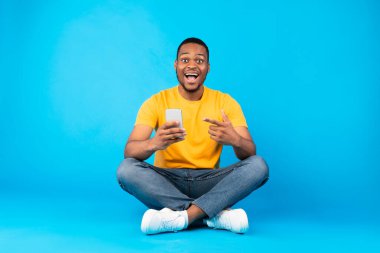 Heyecanlı Afro-Amerikan Akıllı Telefonu Tutan, Stüdyoda Mavi Arkaplanda oturan Heyecanlı Cep Telefonu 'nu işaret eden. Neşeli Adam Cep telefonunuz için Uygulama Önerisi
