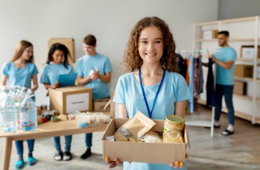 Mavi üniformalı, gülümseyen bir kadın gönüllü bir hayır kurumunda yiyecek bağışlarıyla dolu bir karton kutu tutuyor. Meslektaşları da arka planda bağış düzenlemekle meşgul..
