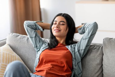 Genç Arap kadın rahat bir şekilde kanepede dinleniyor, elleri başının arkasında, gözleri kapalı, evinde huzurlu bir hafta sonunun tadını çıkarıyor, sakin bir günü kucaklıyor..