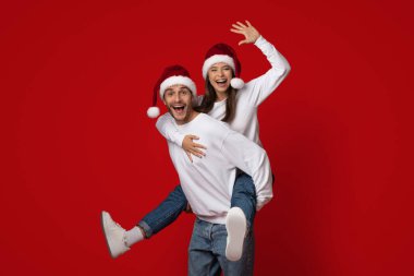 Noel Baba şapkası takan neşeli bir çift birlikte eğlenceli bir anın tadını çıkarır. Adam kadını sırtında taşıyor, her ikisi de sırıtıyor. Canlı kırmızı arka plan şenlikli atmosfere katkı sağlıyor..