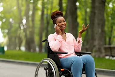 Tekerlekli sandalyedeki neşeli siyahi kadın şehir parkında arkadaşıyla telefon görüşmesi yapıyor. Engelli Afro-Amerikan kadın akıllı telefonuyla birileriyle konuşuyor, dışarıda iletişiminin tadını çıkarıyor.