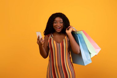 Huge online sale concept. Portrait of excited African American woman with shopping bags using smartphone, purchasing goods on web via new mobile app over orange studio background