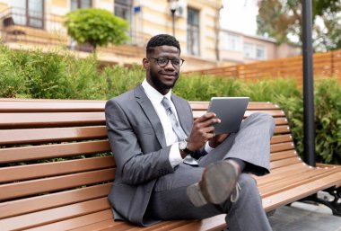 Dijital tablet kullanan kendine güvenen Afrikalı Amerikalı girişimci, şehir merkezindeki modern ofis merkezinin yanındaki bankta otururken haberleri tarıyor.