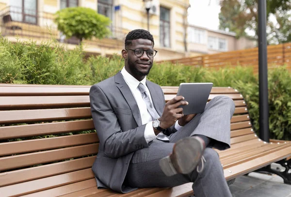 Dijital tablet kullanan kendine güvenen Afrikalı Amerikalı girişimci, şehir merkezindeki modern ofis merkezinin yanındaki bankta otururken haberleri tarıyor.