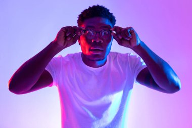 Beyaz gömlekli genç siyah bir adam gözlüklerine dokunuyor, parlak neon ışıkların altında gözleri fal taşı gibi açık. Heyecanlı görünüyor, muhtemelen harika haberlerden ya da büyük bir terfiden bunalmış..