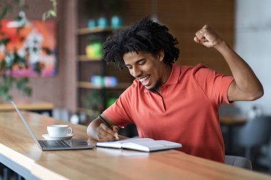 Mutlu, mutlu, heyecanlı, binlerce yıllık Afro-Amerikan kıvırcık adamı zafere seviniyor ve masada bilgisayar ve kafenin içindeki not defteriyle harika sonuçlar elde ediyor. Başarı ve iyi anlaşma, jest ve kazanmak