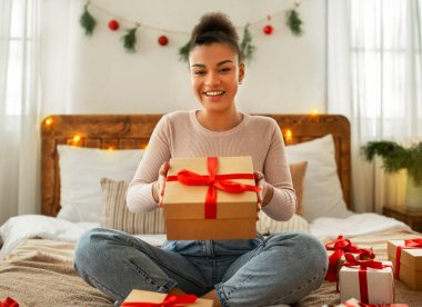 Bu hediye senin için. Afro-Amerikan bayanın elinde kutuyla kameraya veriyor, yatakta oturuyor ve gülümsüyor. Kış tatili kutlaması, yeni yıl teklifi ve Noel hediyeleri.