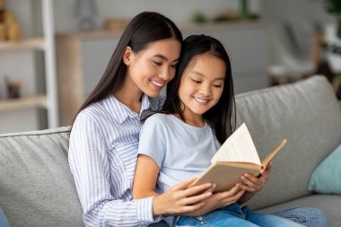 Genç Asyalı bir annenin güzel kızıyla kitap okuması, evdeki kanepede birlikte oturması. Mutlu Koreli anne ve kadın çocuk oturma odasında birlikte vakit geçiriyorlar.