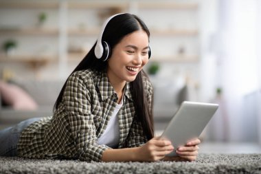 Güzel Asyalı kadın film izliyor, evde yerde yatarken kablosuz kulaklık ve ped kullanıyor, yakın plan, kopyalama alanı. Mutlu genç kadın dijital tablette eğlenceli yeni mobil uygulamalarla eğleniyor