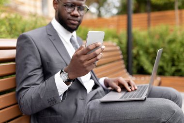 Diz üstü bilgisayarı olan Afrikalı Amerikalı girişimci açık havada bankta oturuyor ve akıllı telefon kullanıyor, seçici odaklanma. Kendine güvenen siyah işadamı müşteriyle telefonda konuşuyor.