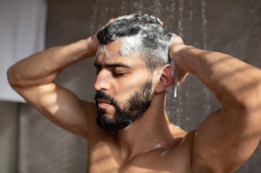 Yakışıklı kaslı adam sıcak bir duş almayı, saçını ve vücudunu şampuanla yıkamayı seviyor. Sıcak suyun altında duruyor, köpükle kaplıyor ve evinde parlak bir banyo setiyle onu örtüyor..