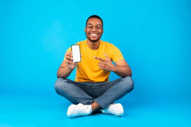 Gülümseyen Afrikalı adam cep telefonunu boş ekranla gösteriyor. Mavi Stüdyo Arkaplan 'da oturan akıllı telefon için harika bir uygulama öneriyor. People and Gadgets, Yeni Başvuru. Model